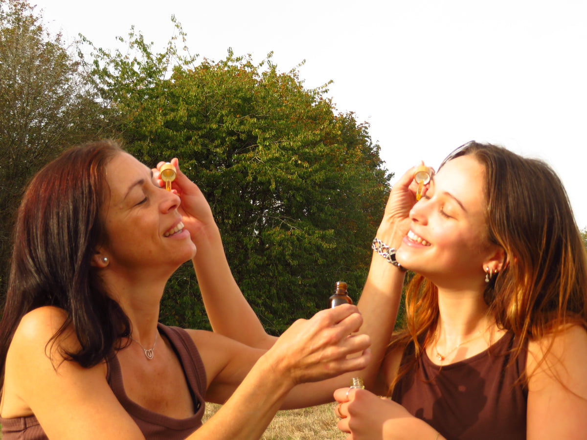Two women outdoors applying a small cosmetic or beauty product to their faces, with trees in the background.