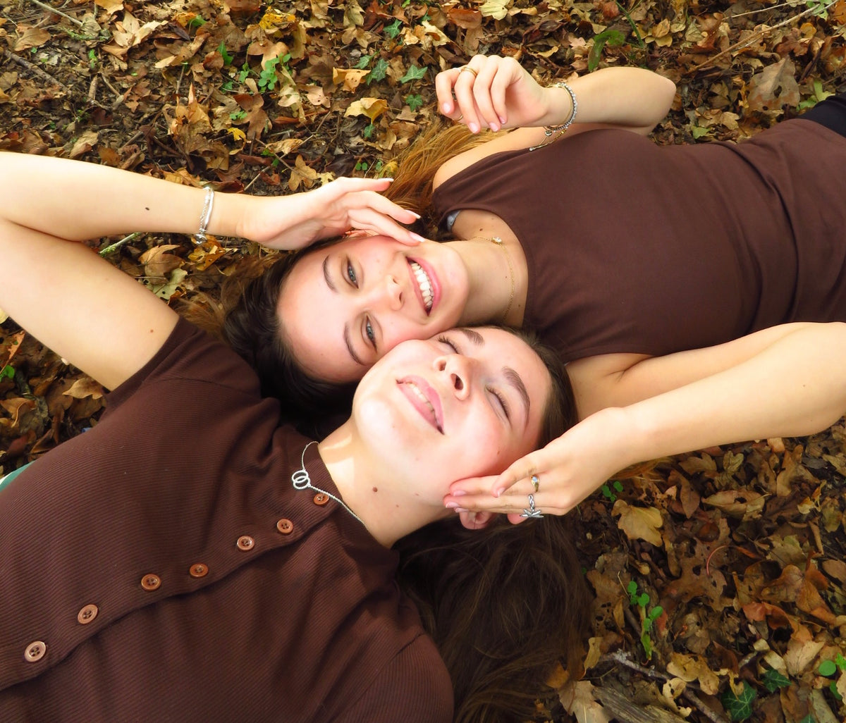 Two women lying on a bed of fallen autumn leaves, wearing matching brown button-up shirts and looking up at the camera with smiles.