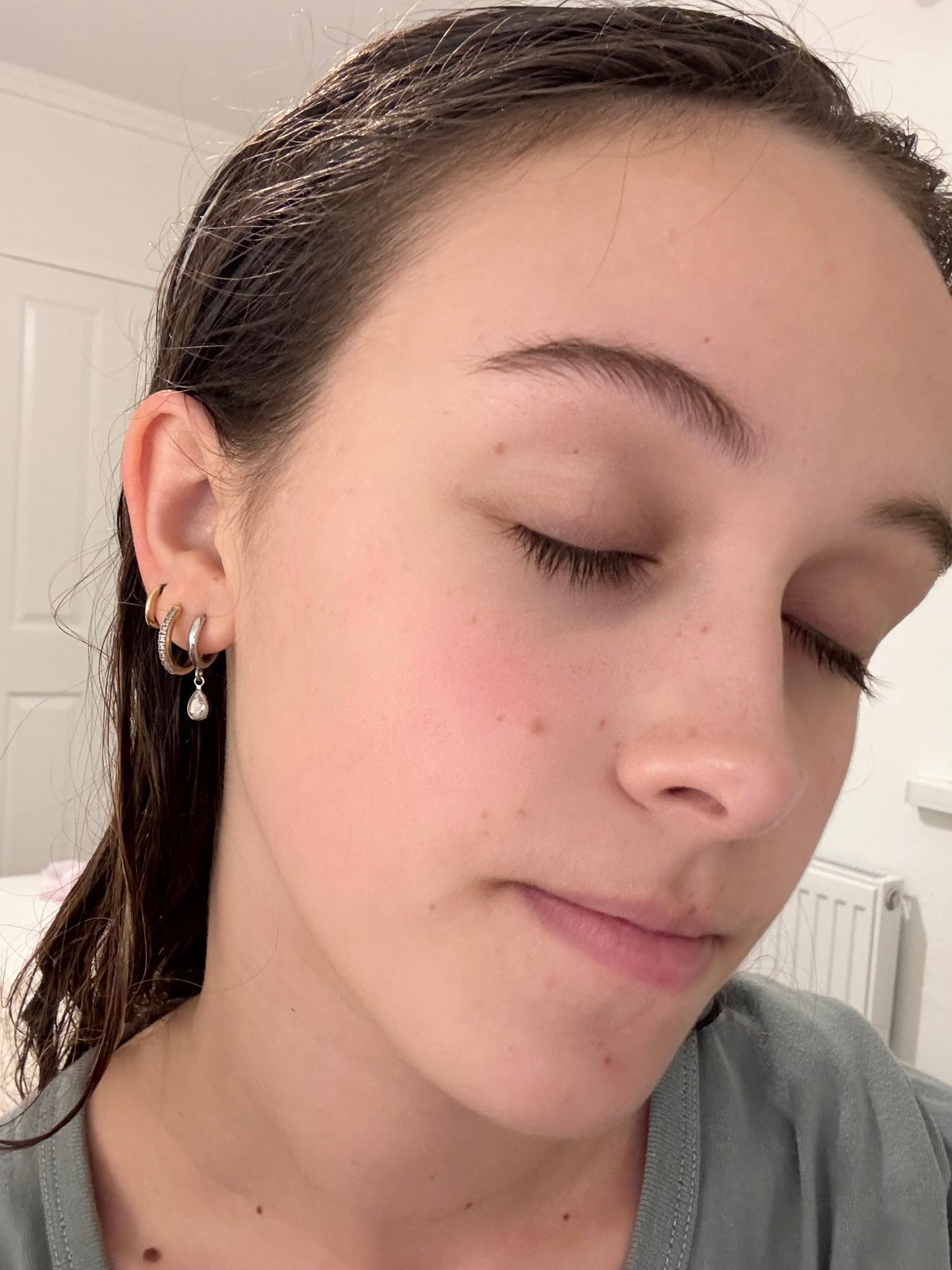 A close-up shot of a person wearing small gold hoop earrings with a dangling pearl-like bead on one earring, shown in a casual indoor setting.
